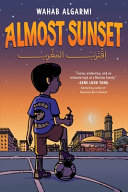 Boy standing on a soccer field with the ball beneath his foot looking out at the sunset over the buildings.